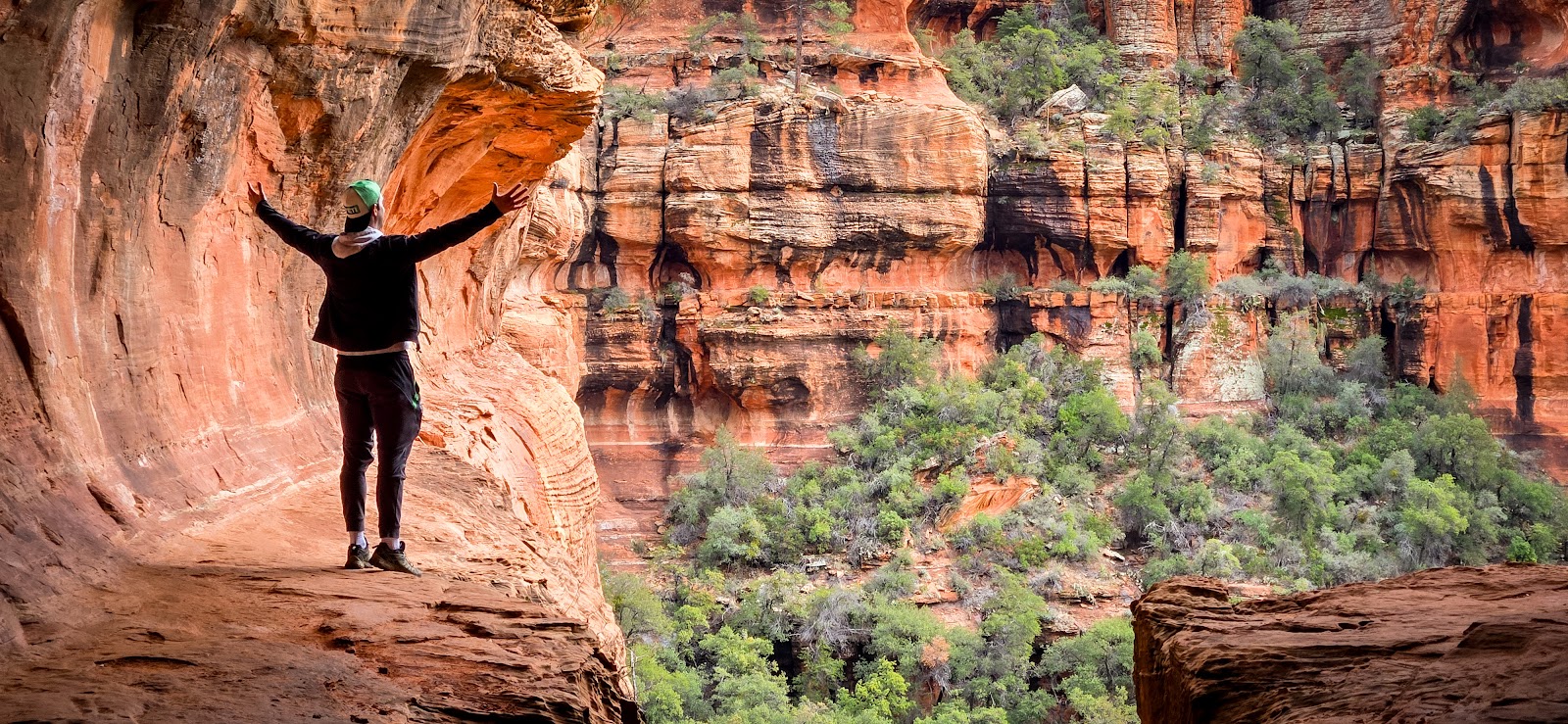 Person standing on a red rock ledge with arms raised above a Sedona canyon